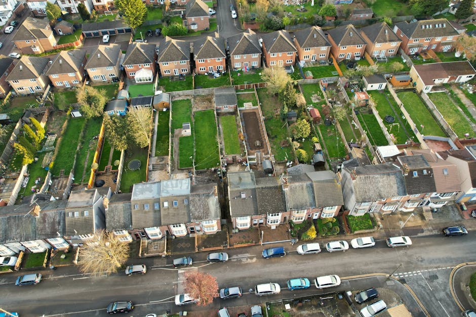 An aerial view of a residential neighborhood showing a row of terraced houses with front gardens and driveways. The backyards are visible, featuring well-maintained lawns, garden beds, paved pathways,