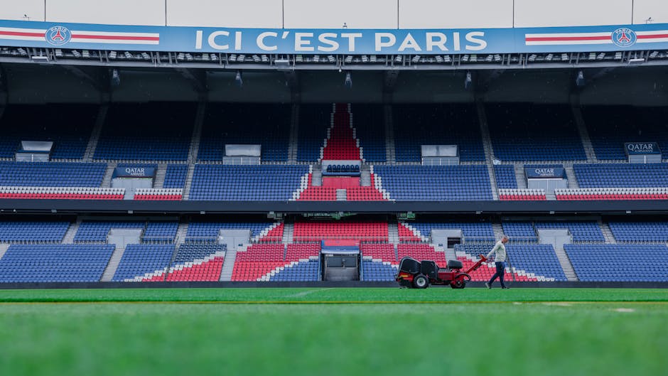 A gardener from Gardeners Selhurst is seen maintaining a sports stadium's grass field, using a walk-behind lawn mower to trim the lush, green grass. The stadium seating is visible in the background, f