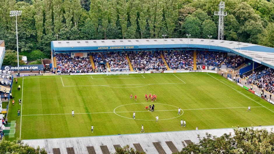 A gardener from Gardeners Selhurst is seen maintaining a sports stadium's grass field, using a walk-behind lawn mower to trim the lush, green grass. The stadium seating is visible in the background, f