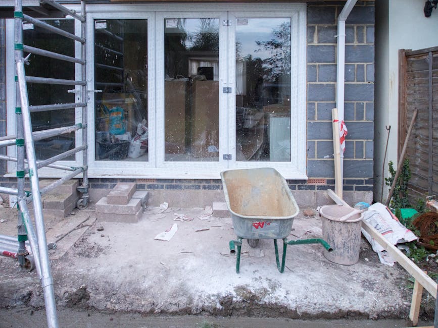 A rear garden area under renovation, with construction tools and materials scattered across the space. The scene features a partially finished patio with uneven, dusty ground and a wheelbarrow positio
