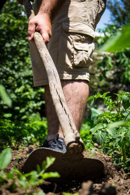 A rear garden area under renovation, with construction tools and materials scattered across the space. The scene features a partially finished patio with uneven, dusty ground and a wheelbarrow positio